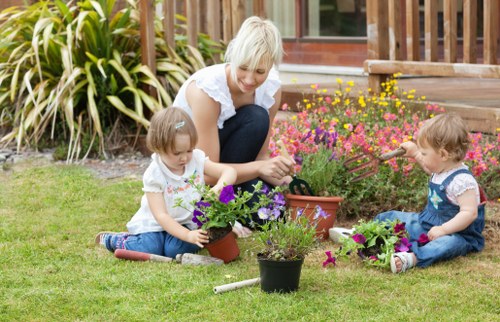 A woman with short blonde hair, dressed in a white top, is sitting on a well-maintained lawn in a garden in Hornsey, engaging with two young children who are planting and tending to potted flowering plants. The children, one girl with light brown hair and another with darker hair, are sitting cross-legged on the grass, each handling small plant pots containing vibrant purple and pink flowers. The garden features lush, green grass, with flower beds filled with a variety of colourful blooming plants, including pink and yellow flowers, situated near a wooden deck with stairs leading up to a house. Tall, leafy plants with variegated foliage border the garden's background, providing a natural screen. The scene is lit by natural daylight, suggesting a clear, pleasant day. This outdoor space exemplifies a tidy, landscaped garden suitable for family outdoor activities and is representative of the type of garden care and landscaping services offered by Lawn Mowing Hornsey to maintain lush, inviting garden environments in the Hornsey area.