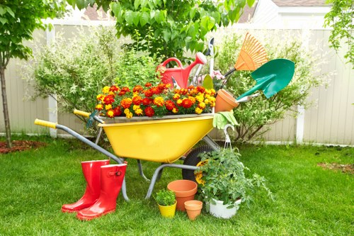 A colourful garden scene in a residential outdoor space with a lawn area featuring well-maintained, lush green grass. The foreground displays a yellow wheelbarrow filled with vibrant red, orange, and yellow flowering plants, positioned on a neatly kept lawn. Surrounding the wheelbarrow are gardening tools and accessories, including a pair of red rubber boots placed on the grass, along with terracotta plant pots and a small white container holding green foliage. In the background, there are leafy trees and shrubs providing natural shade and privacy, with a white garden fence visible behind them. The overall setting appears to be in Hornsey, with a bright, sunny day enhancing the natural tones of the grass, plants, and garden elements, reflecting the professional gardening and lawn maintenance services offered by Lawn Mowing Hornsey.