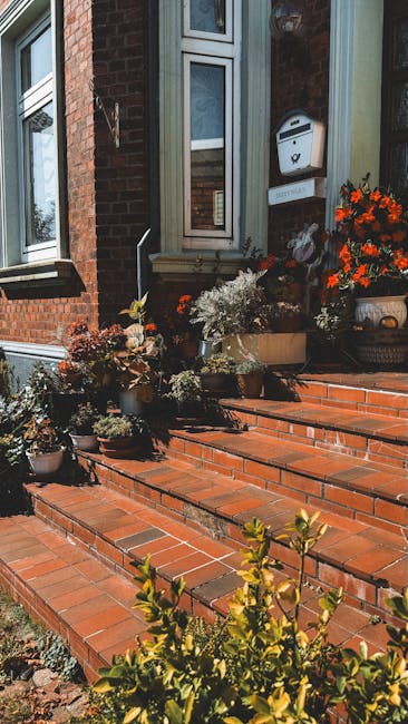A close-up view of a red-brick residential building's front garden area, featuring a large, leafy shrub with green and reddish-orange foliage growing against the exterior wall. The house has white-fra